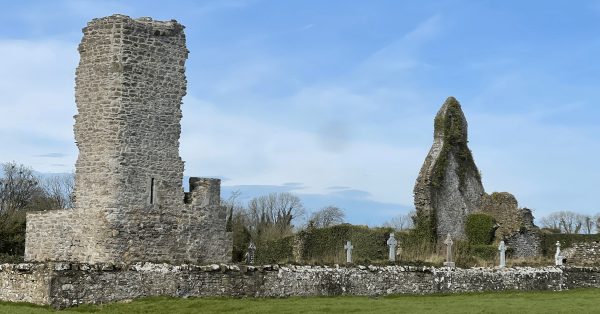 Ruins of an old stone church and tower stand in a grassy area, surrounded by a stone wall and visible gravestones, with trees and blue sky in the background.