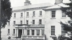 Black and white photo of a large, historic, three-storey building with many windows, a main entrance with columns, and trees partially framing the view.
