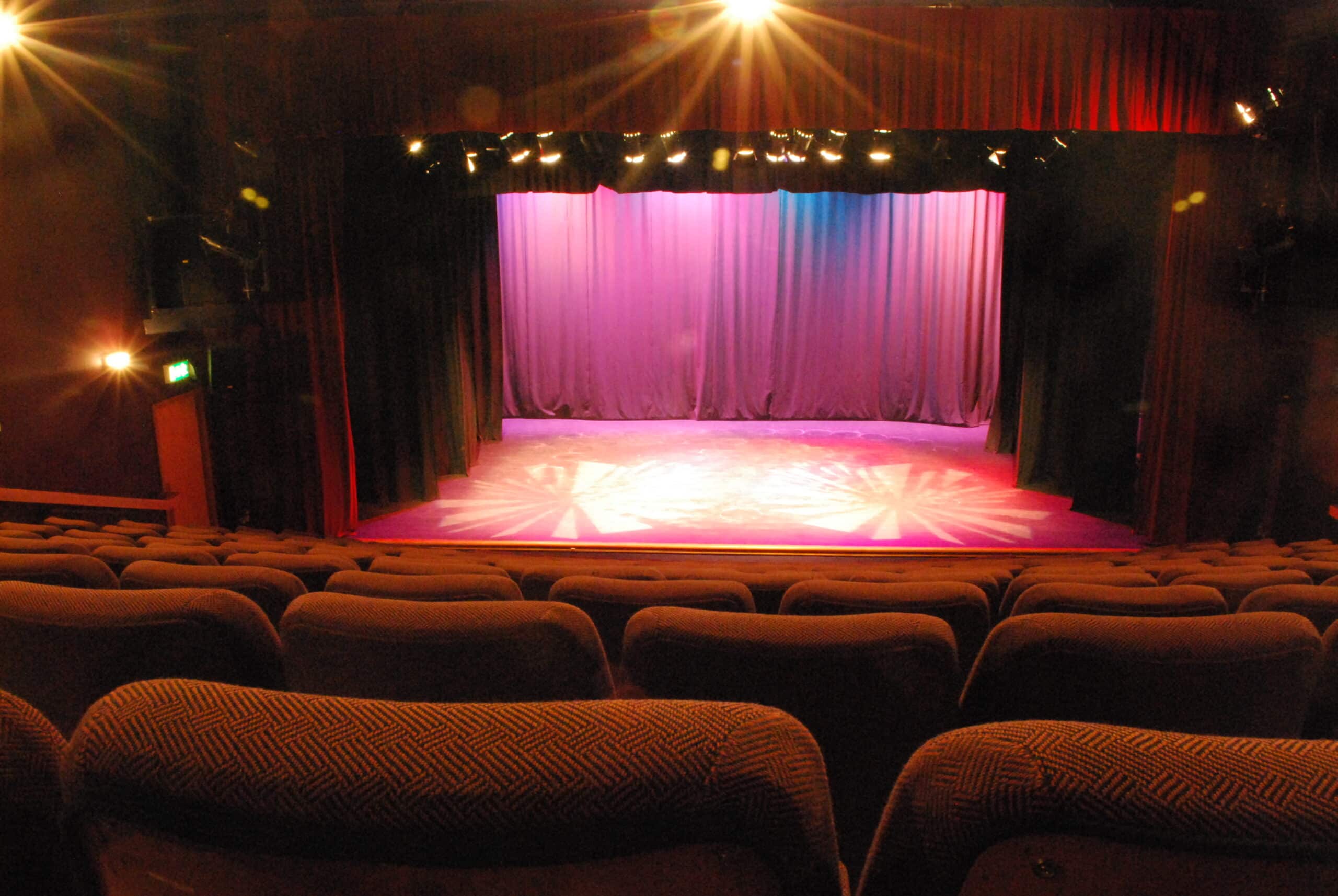 Rows of empty theatre seats face a lit stage with purple curtains and spotlights, creating a dramatic ambience.