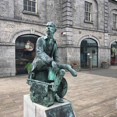 Bronze statue of a seated man with papers on his lap, positioned in front of a grey stone building with arched windows and doors.