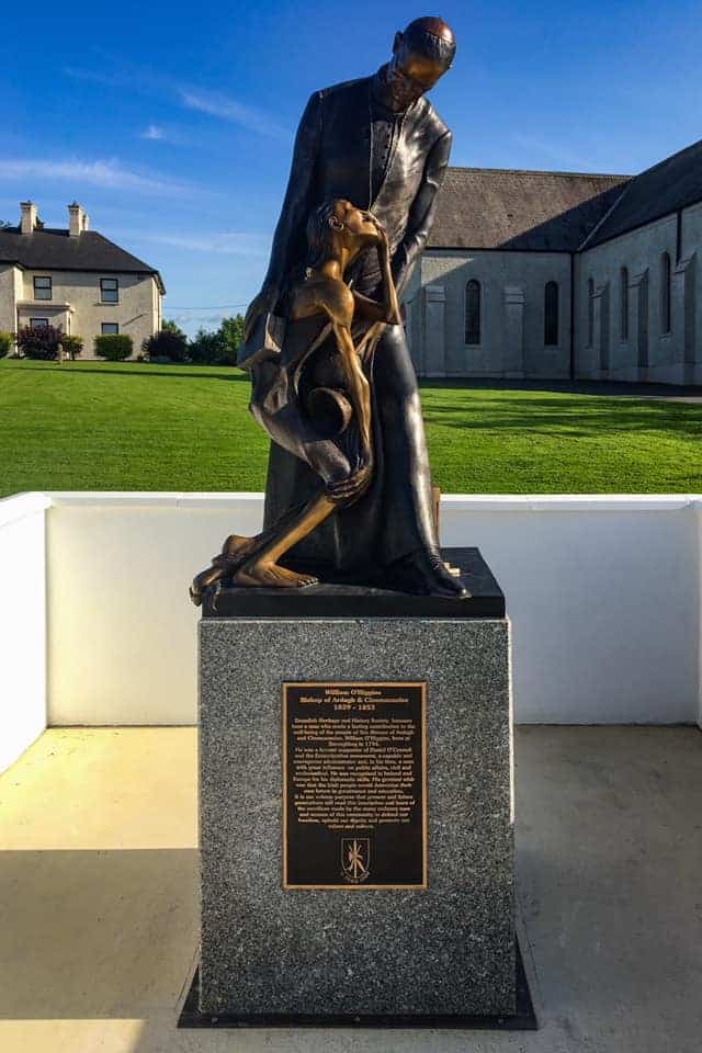 A bronze statue of a man holding a weak, emaciated person on a stone plinth with a plaque, set outdoors near grass and buildings under a blue sky.