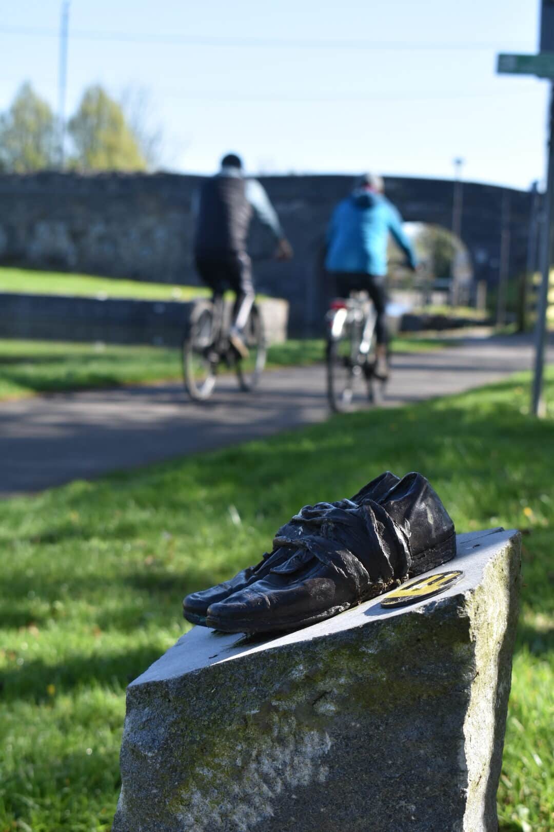 A black cycling shoe sculpture is mounted on a stone plinth in the foreground, with two cyclists riding on a path and a stone bridge visible in the blurred background.