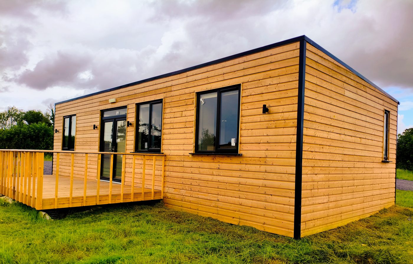 A modern, single-storey wooden cabin with black-trimmed windows and a small deck sits on a grassy field under a cloudy sky.