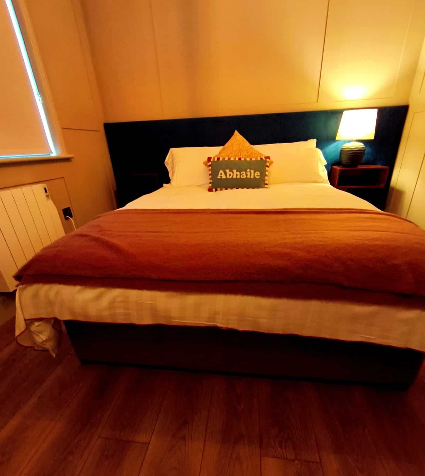 A cosy bedroom with a double bed, white and rust-coloured bedding, a pillow labelled "Abhaile," a lit bedside lamp, wooden floor, and a window with the blinds partially closed.