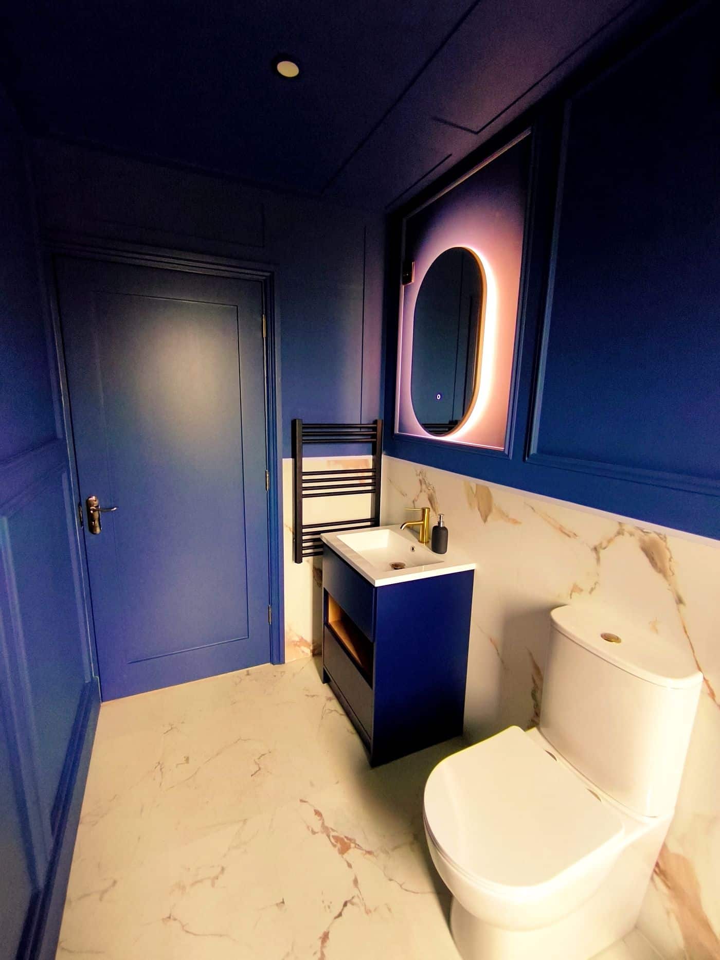 A modern bathroom with blue walls, a round LED-lit mirror above a basin, a white toilet, marble-patterned flooring, and a heated towel rail beside the blue door.