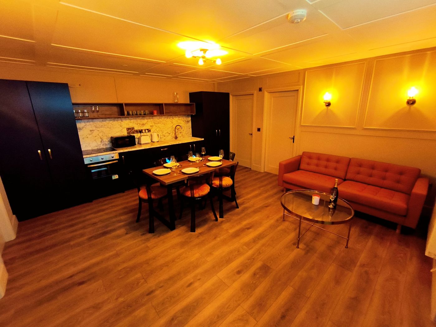 A cosy kitchen and living area with wooden floors, orange sofa, glass coffee table, dining table set for four, and dark cupboards against a wall with marble splashback. Warm yellow lighting fills the room.