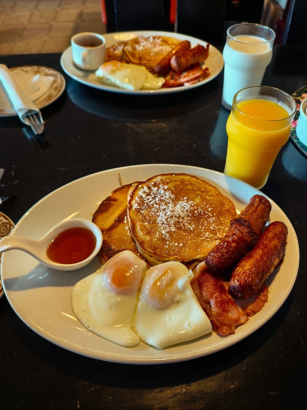 Two plates of breakfast with pancakes, syrup, fried eggs, sausages, and bacon. There are glasses of milk and orange juice, plus a cup of coffee on a dark table.