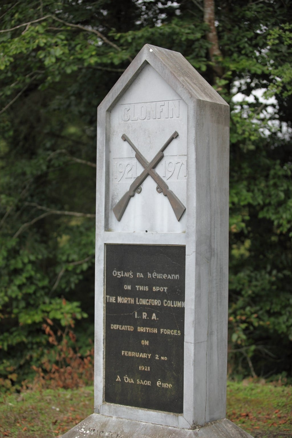 Gravestone-shaped monument with crossed rifles, dates 1921–1971, and an inscription commemorating the North Longford Column defeating British forces, surrounded by trees in the background.
