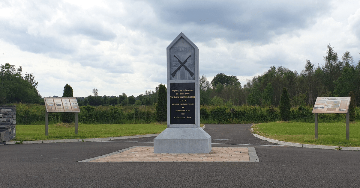 A stone memorial with plaques stands on a paved area by a road, surrounded by grass and trees. Two informational signs are on either side, and the sky is cloudy.