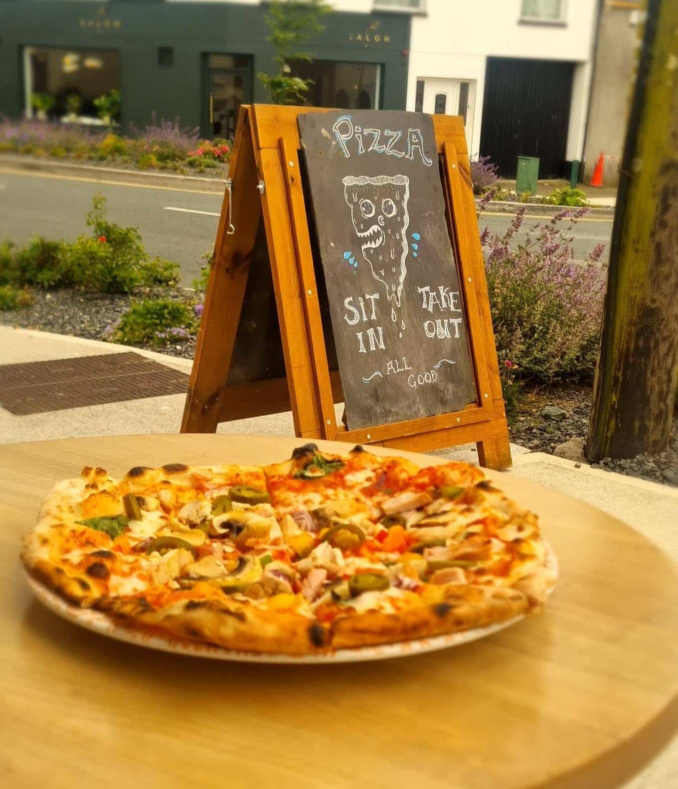 A plate of pizza on a table outside, with a chalkboard sign in the background that reads, "Pizza, Eat In, Takeaway, All Good," and a pizza slice drawing.