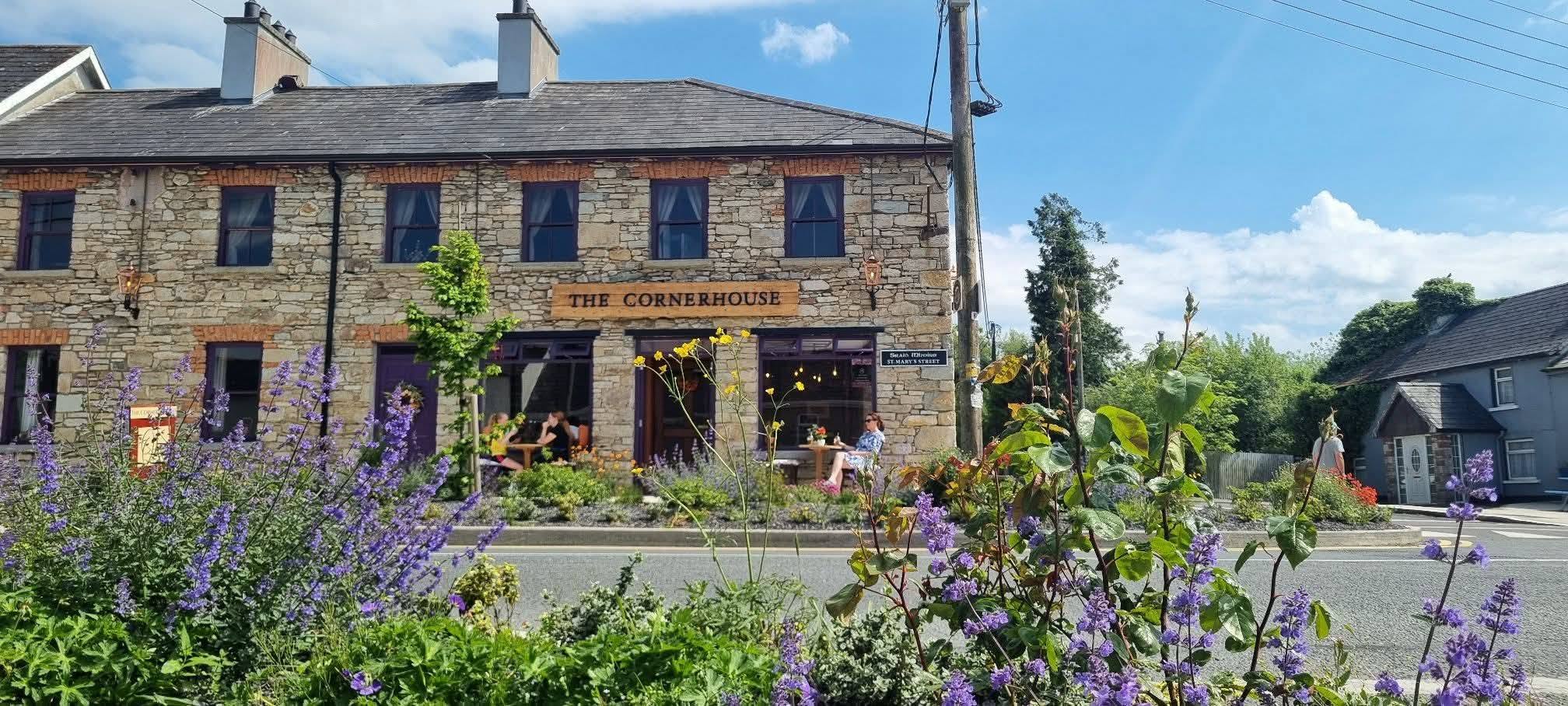 A stone building labelled "The Cornerhouse" sits across the street, with people at outdoor tables and purple flowers in the foreground under a bright blue sky.
