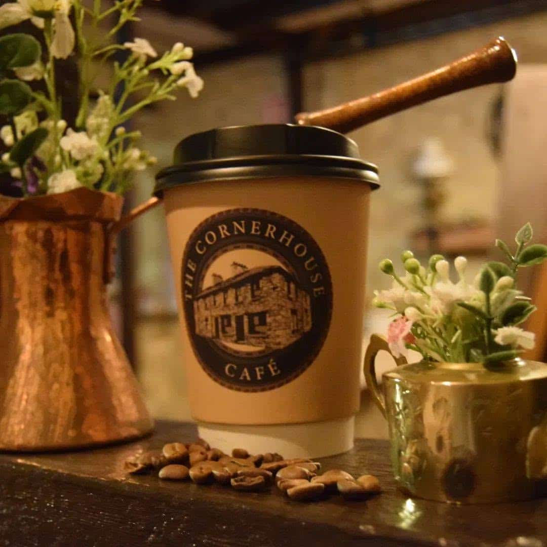 A coffee cup labelled "The Cornerhouse Café" sits on a wooden surface, surrounded by flowers in copper containers and scattered coffee beans.