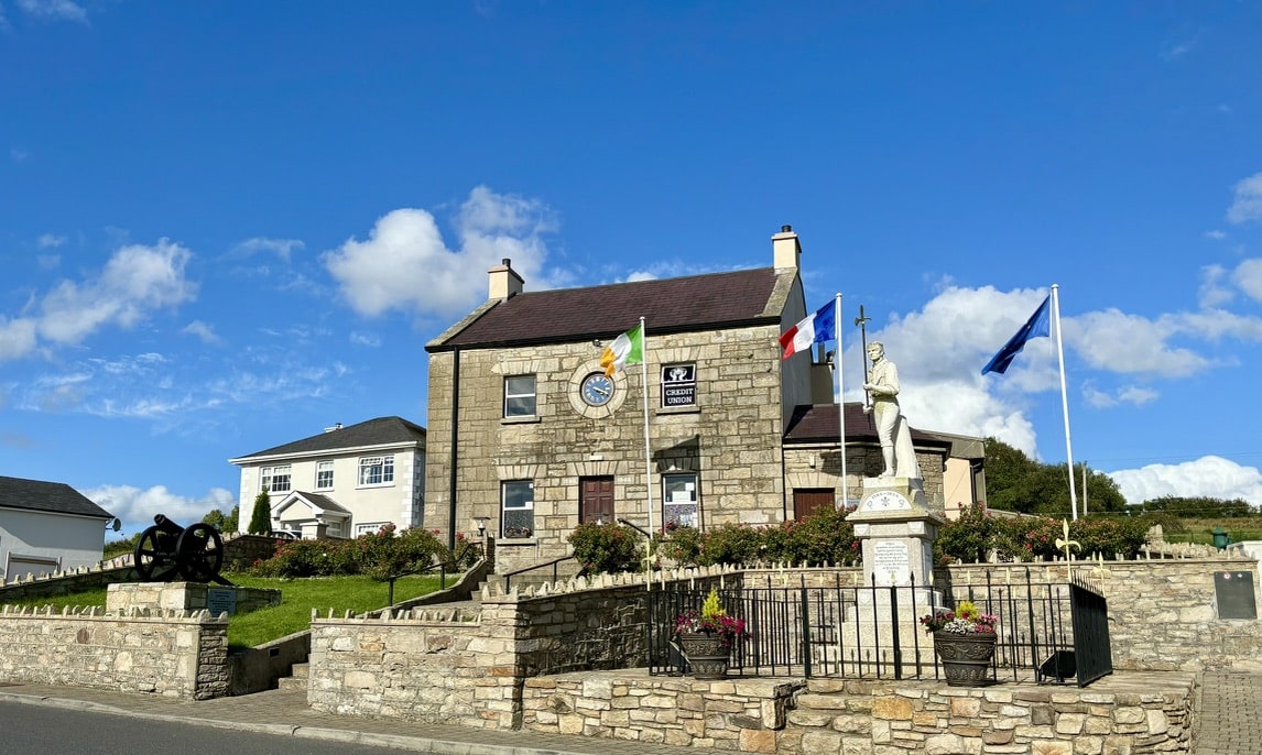 Stone building with a clock, several flags, a white statue, flowerbeds, and a cannon in front, all under a bright blue sky with scattered clouds. The area is fenced with low stone walls.