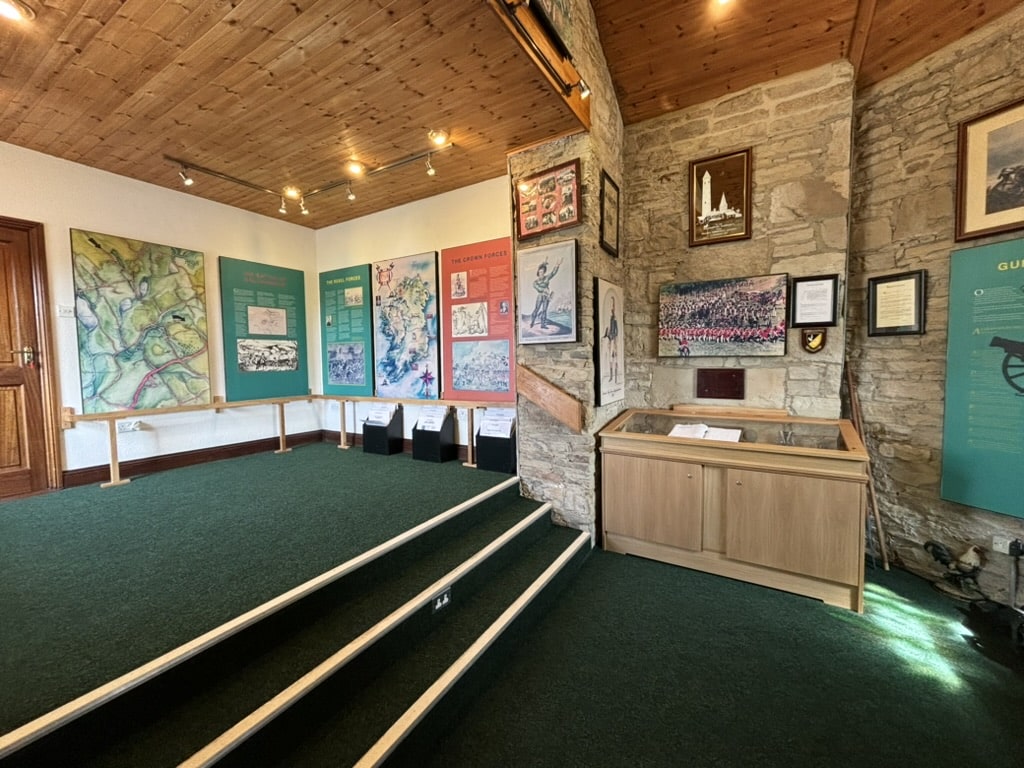 A small museum room with green carpet, stone walls, display boards with maps and photos, framed pictures, and a glass display case. Wooden ceiling and steps lead up to the exhibits.