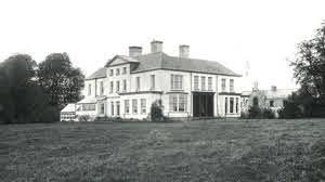 Black and white photo of a large, two-storey stately home with multiple chimneys, surrounded by grass and trees, with a smaller building visible to the right.