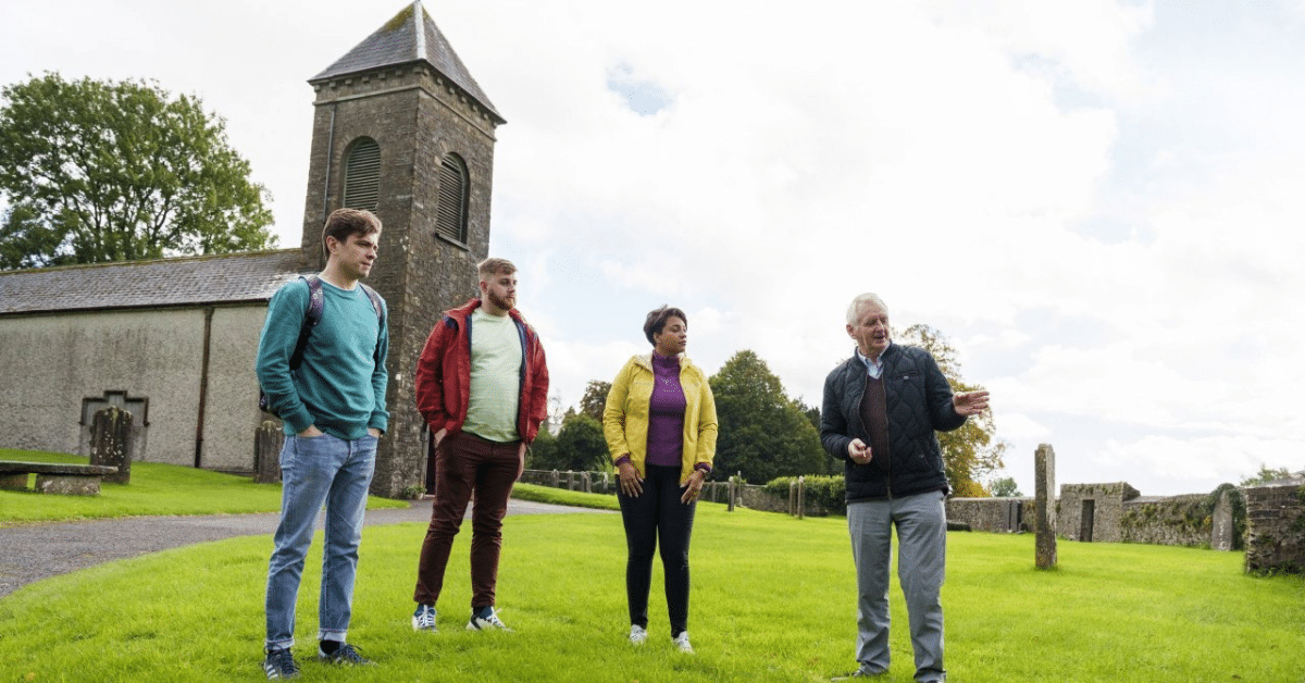 A guide speaks to three people standing on grass near an old stone church with a tower. The group listen attentively under a cloudy sky, surrounded by green trees and gravestones.