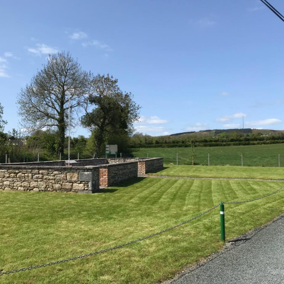 A neatly mown lawn bordered by a stone wall sits beside a country road. Trees and a green field are in the background under a clear blue sky.