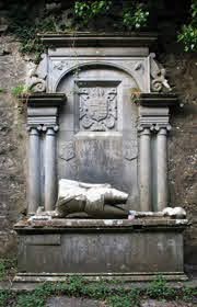 Weathered stone monument with columns and decorative carvings, featuring a broken statue lying on the base, set against an old stone wall with some creeping greenery above.
