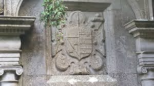 Stone relief of a worn coat of arms carved into a building façade, partially covered by a small green plant hanging above.