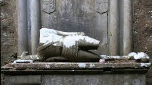 A weathered stone monument displays only a pair of legs in armour, resting on a platform, partially covered with snow against a stone wall.