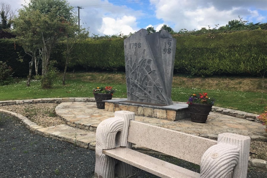 A stone monument engraved with "1798" and "1998" stands in a circular paved area, flanked by potted flowers, with a white stone bench in the foreground and green hedges and trees in the background.