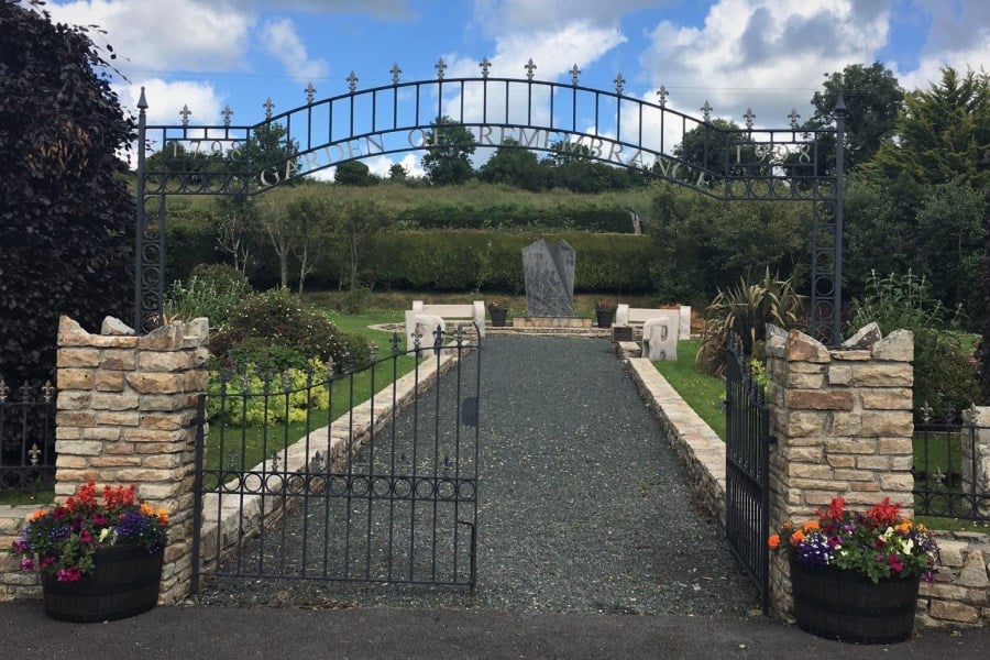 An open iron gate reading "Garden of Remembrance" leads to a gravel path flanked by stone walls, benches, and two stone memorials, with colourful flowers in planters and greenery in the background.