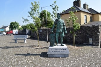 A statue of General Sean MacEoin stands on a stone plinth beside two small trees in a paved square, with a yellow building, stone wall, and bench visible in the background.