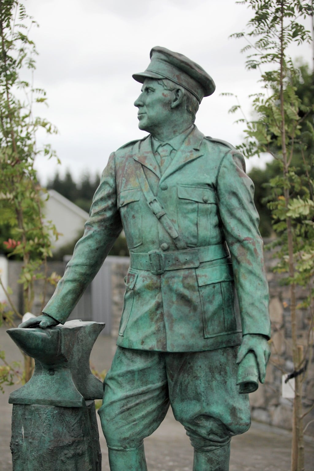 A bronze statue of a man in a military uniform and cap stands outdoors, holding an object near a stone wall and trees.