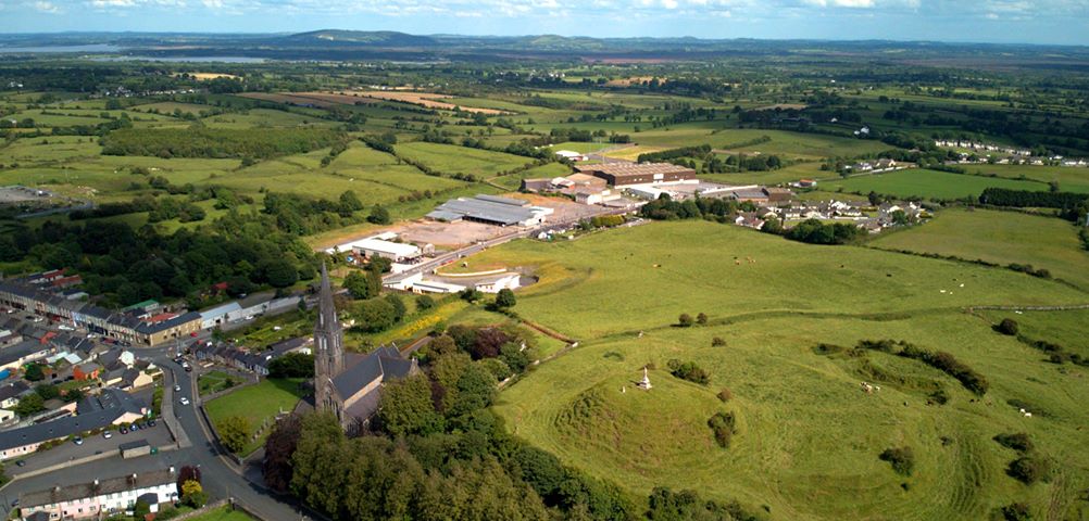 Aerial view of a rural landscape with fields, a church with a tall spire, scattered buildings, and rolling hills under a partly cloudy sky.