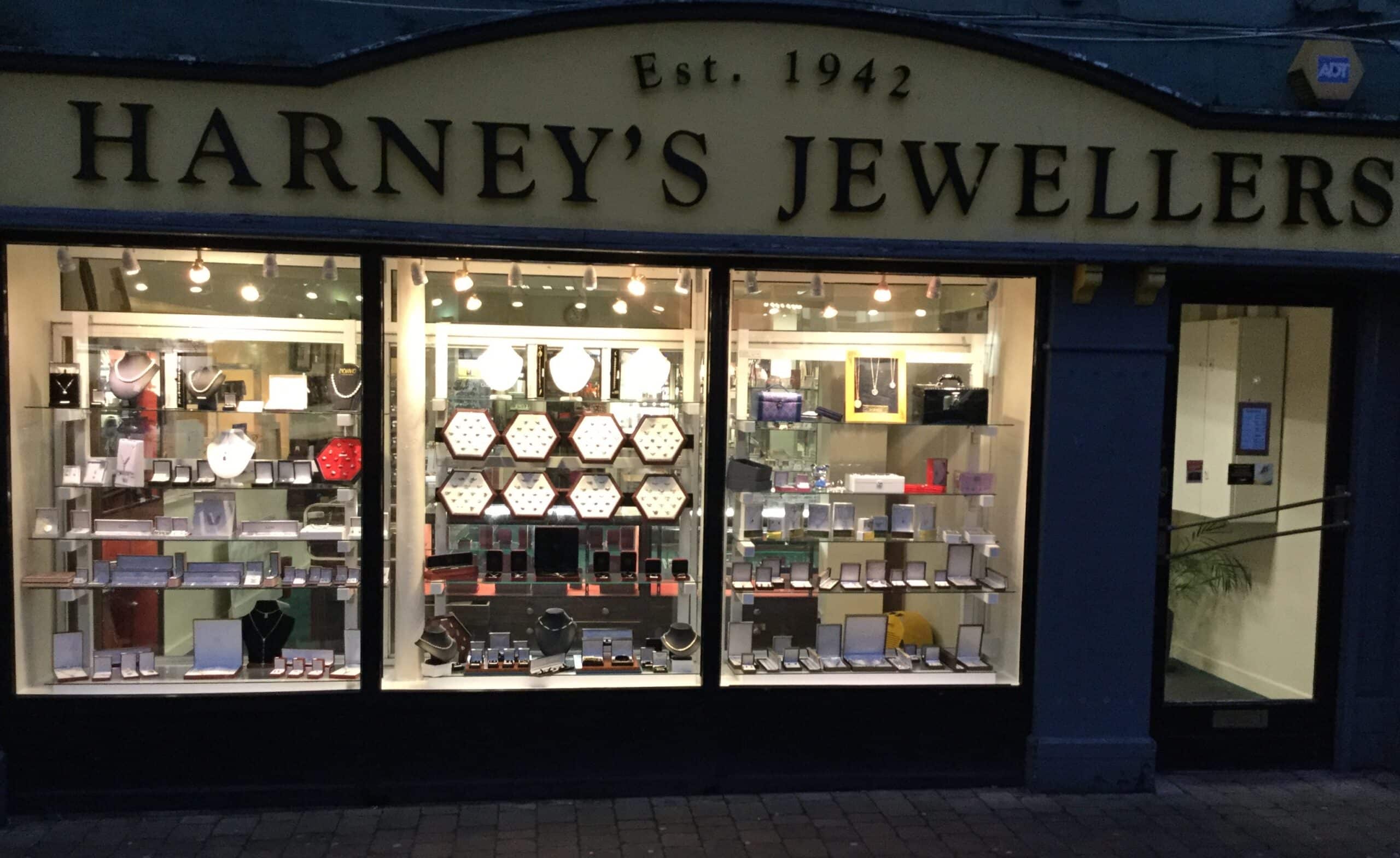 Shopfront of Harney’s Jewellers, established 1942, with window displays of various jewellery pieces like necklaces, watches, and rings, illuminated by warm lights on a brick walkway in early evening.