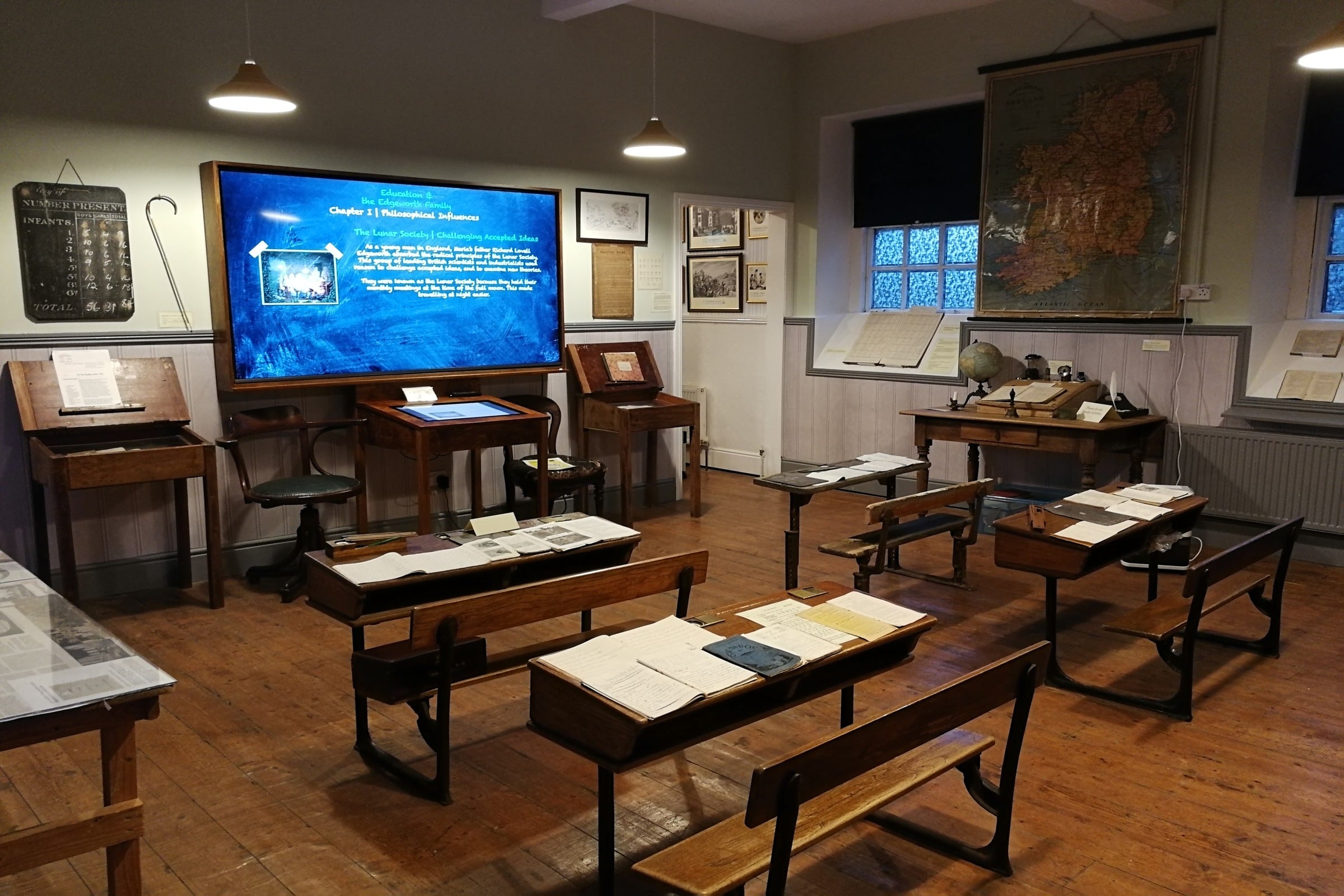A historical classroom display with wooden desks, benches, old books, maps, and a digital screen showing educational content, evoking an early 20th-century school atmosphere.