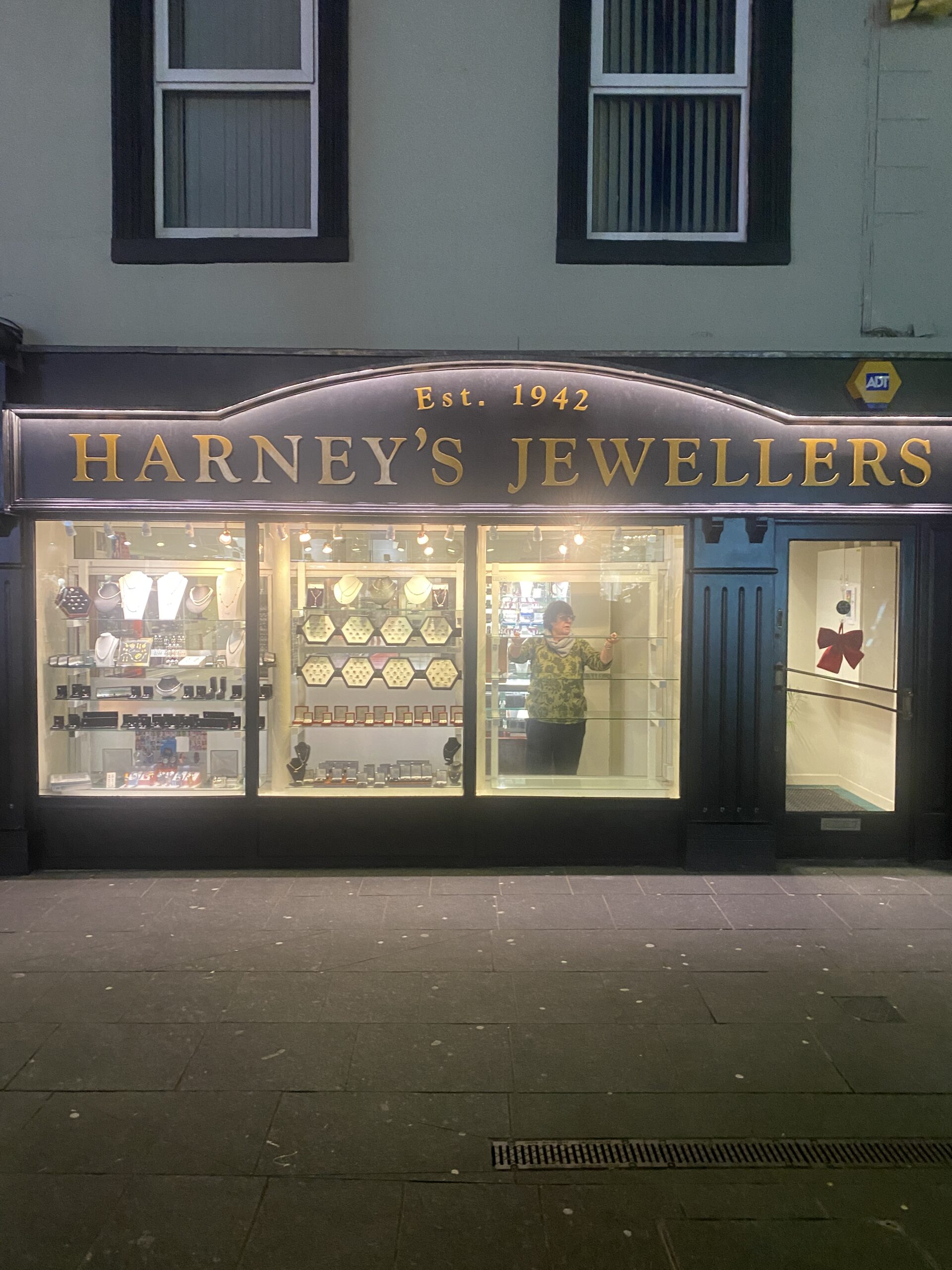 A jewellery shop called "Harney's Jewellers" with a display of necklaces and watches in the window. A person stands inside the brightly lit shop, visible through the glass. The sign says "Est. 1942.
