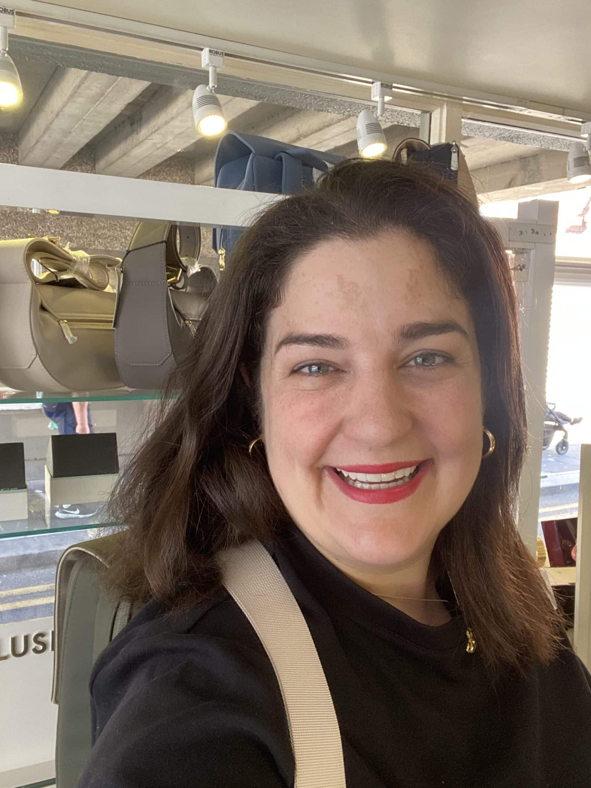 A woman with dark hair and red lipstick smiles at the camera inside a shop with handbags displayed on shelves behind her.