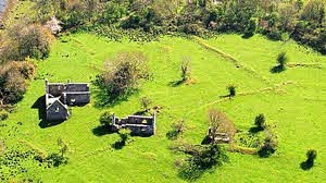 Aerial view of a green field with scattered trees and three old, partially ruined stone buildings surrounded by grass. Some paths cross the landscape.