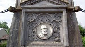 Stone monument featuring a circular carved portrait of a bearded man, surrounded by ornate decorative details and patterns, set into a triangular pediment.