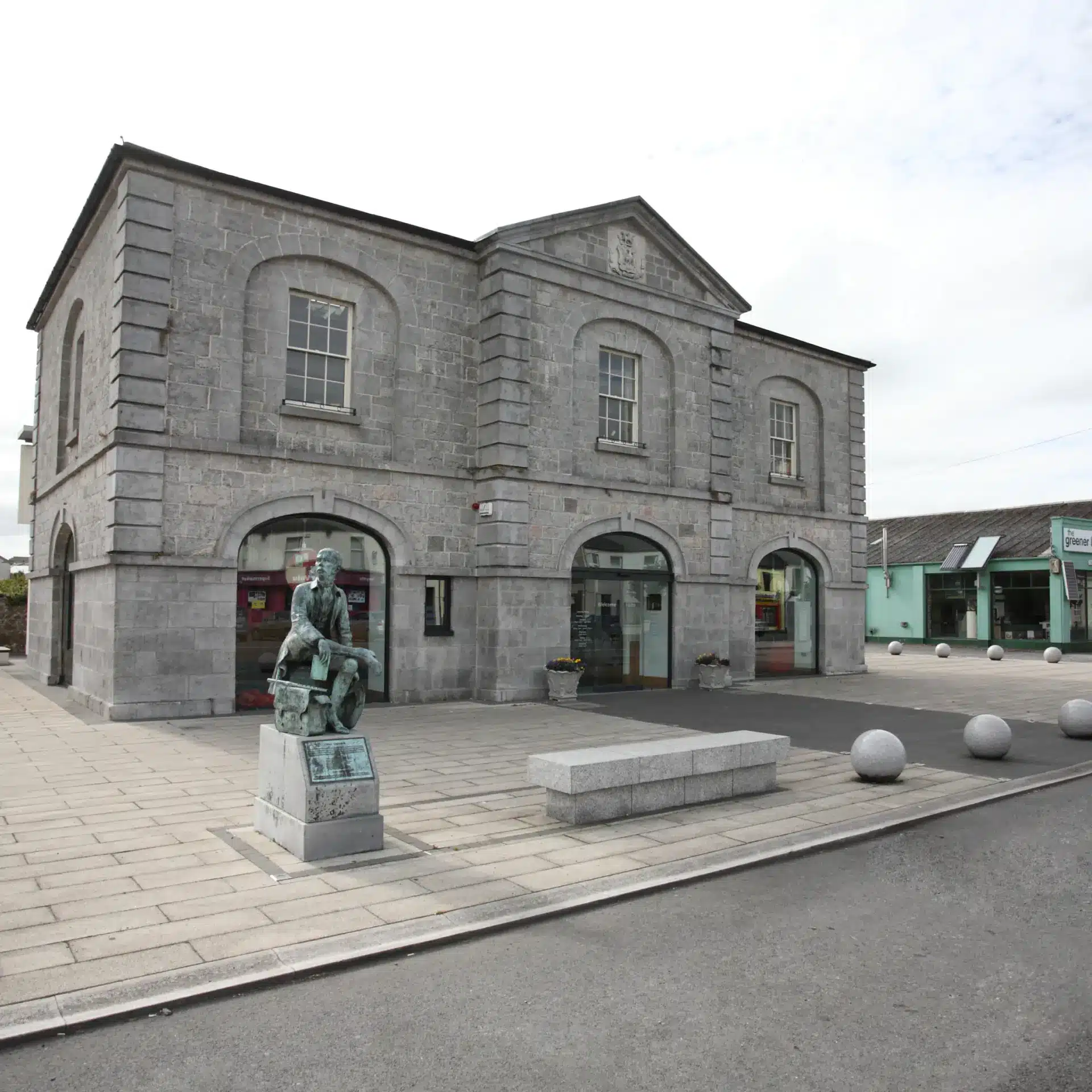A grey stone building with arched doorways and windows sits on a paved square. In front stands a bronze statue of a seated person on a plinth. Nearby are a stone bench and round concrete bollards.