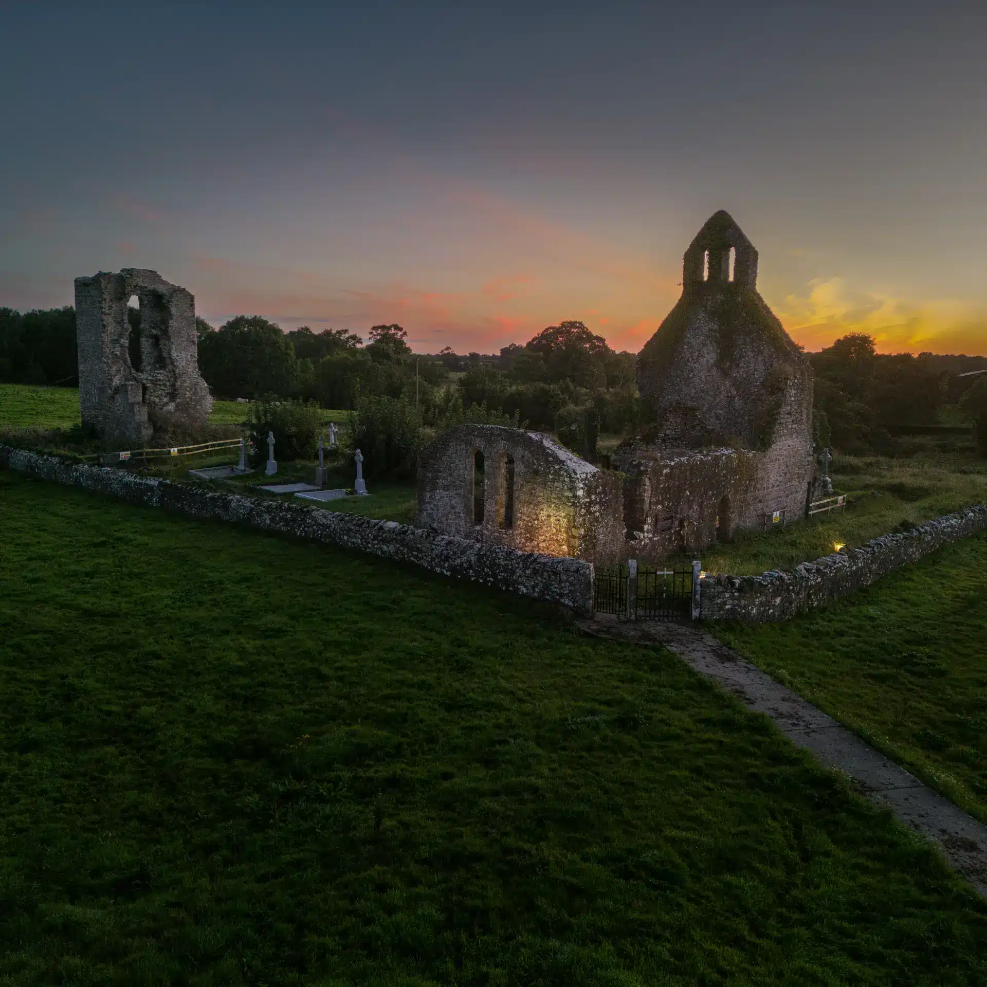 An aerial view of ancient stone ruins and grave markers surrounded by a stone wall, set against a grassy landscape at sunset with a colourful sky in the background.