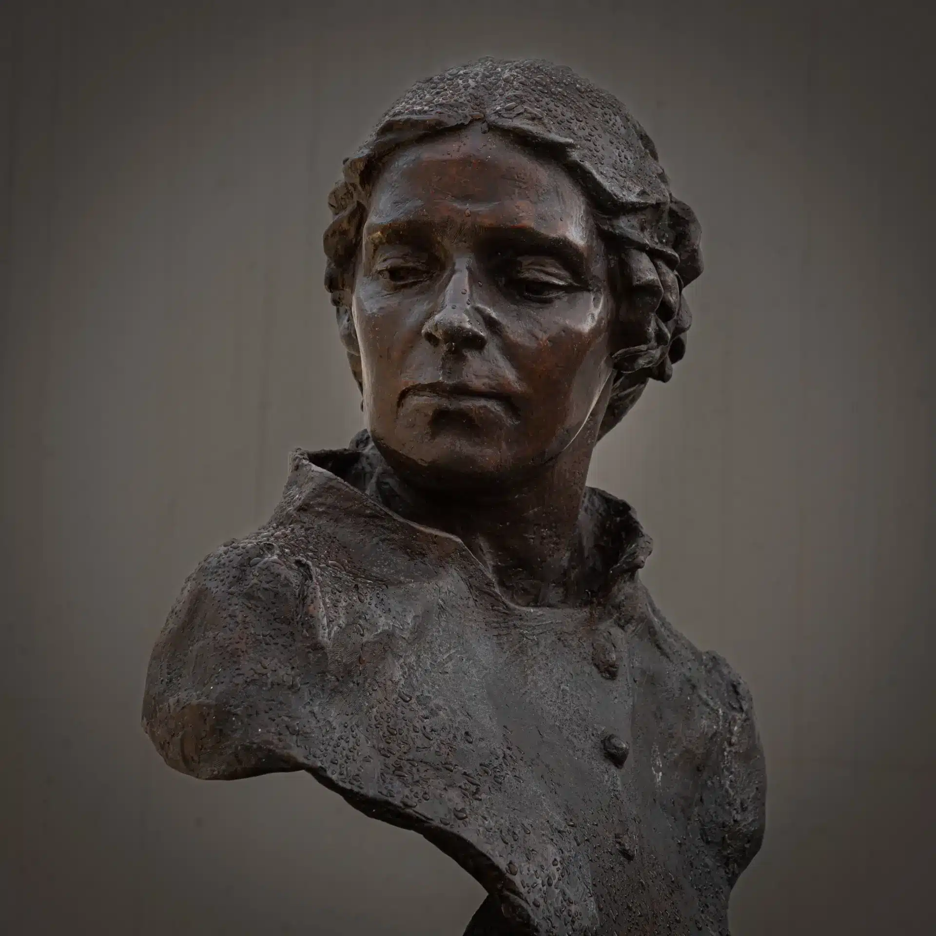 A bronze bust sculpture of a woman with a solemn expression, wearing a collared garment, set against a plain, softly lit background.