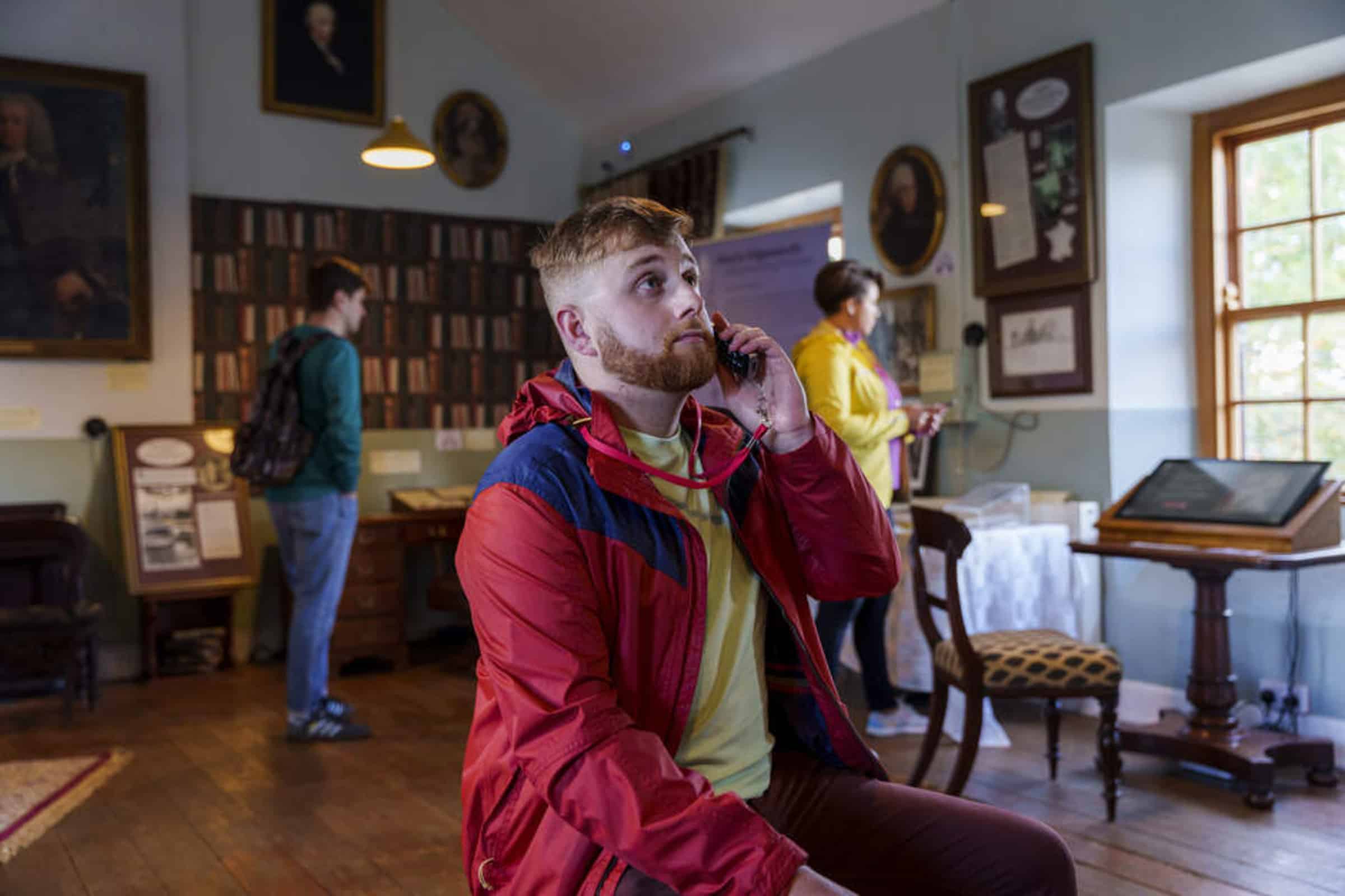 A man in a red jacket listens to an audio guide in a museum room, whilst two other visitors explore exhibits in the background. Paintings and framed pictures hang on the walls.