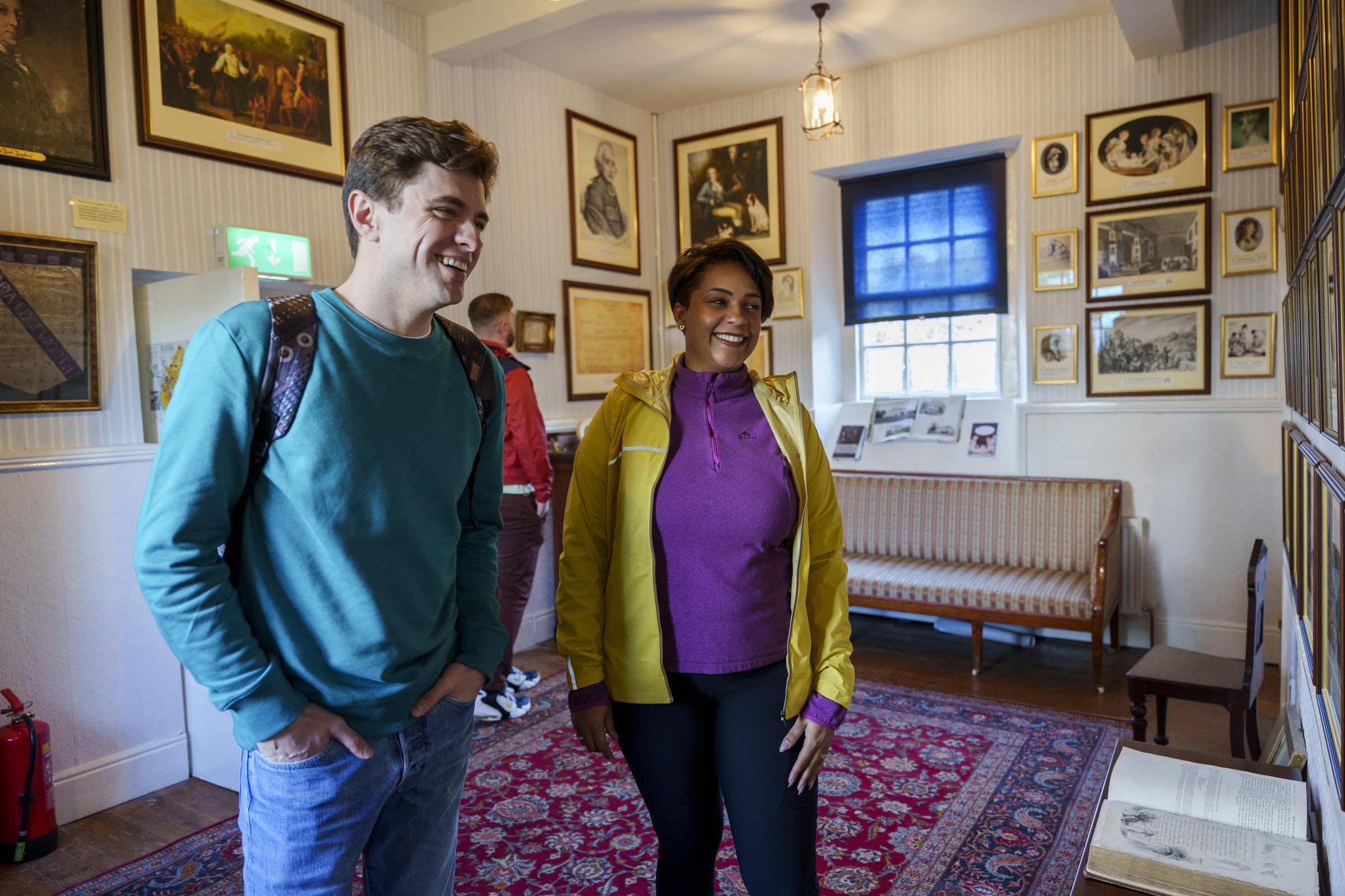 Two people stand and smile whilst viewing framed pictures and documents on a wall inside a cosy, well-lit room decorated with art, a red patterned rug, a bench, and historical photographs.