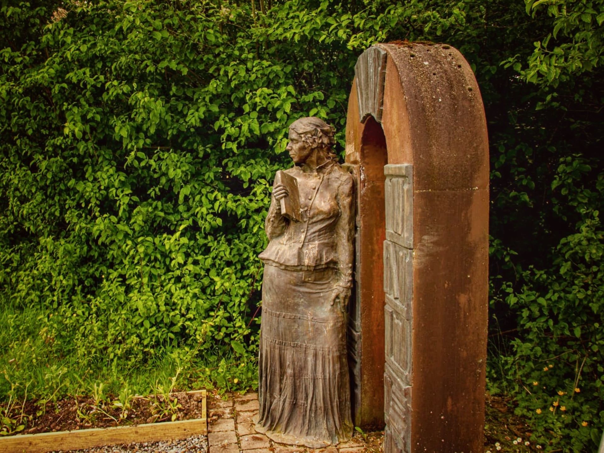 A bronze statue of a woman in historical clothing stands beside an arched doorway, surrounded by lush green foliage and a cobbled path.