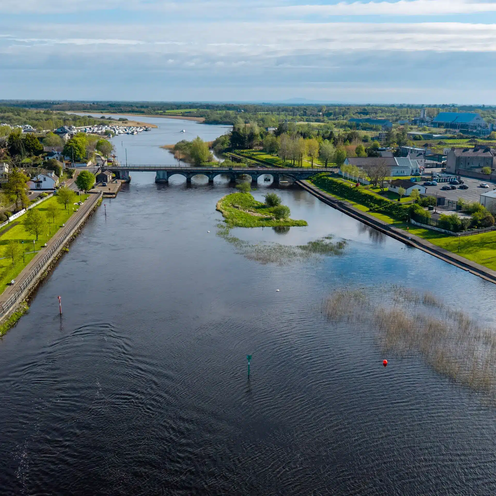 Aerial view of a river with a small bridge, grassy banks, scattered trees, and a nearby village under a partly cloudy sky. In the background, boats are moored near the river’s mouth.