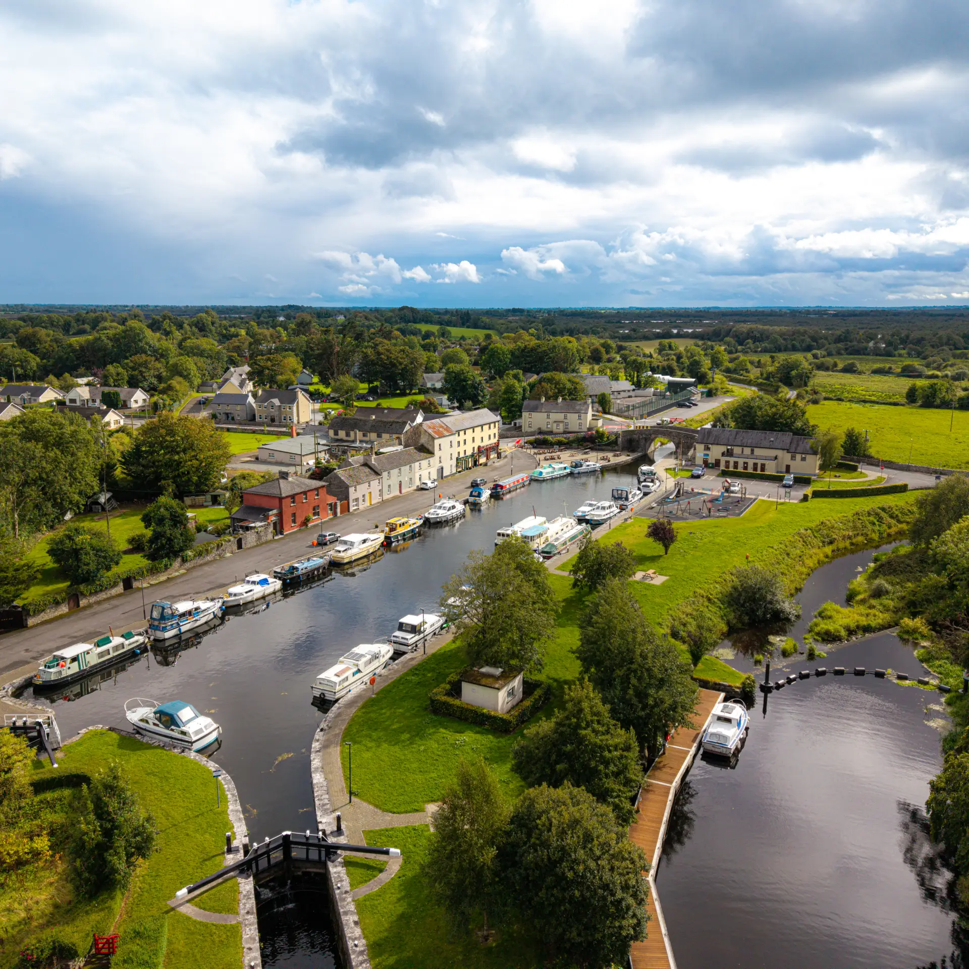 Aerial view of a small town with boats moored along a winding river, surrounded by green fields, trees, houses, and a partly cloudy sky.