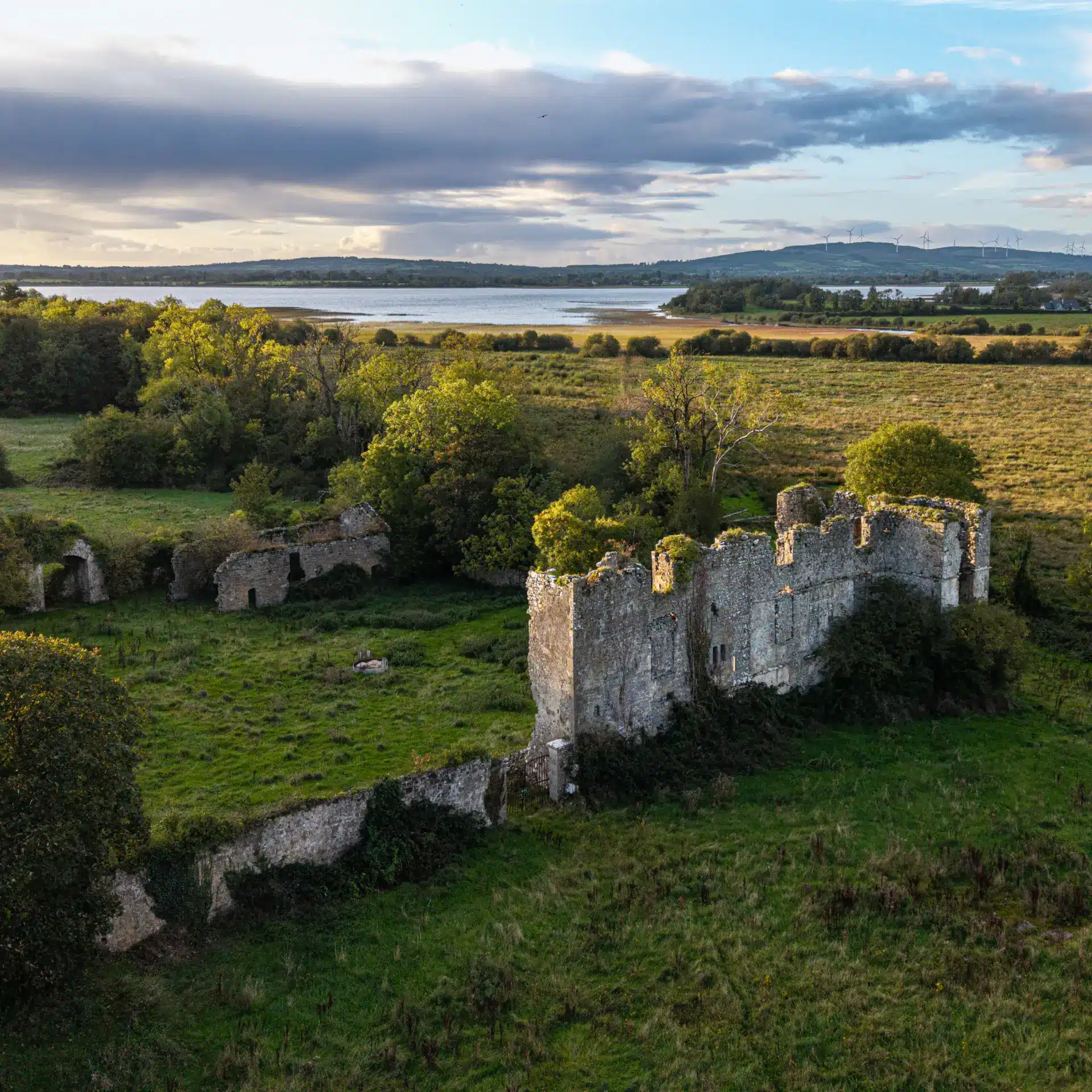 Aerial view of an old stone castle ruin surrounded by green fields and trees, with a lake and rolling hills in the background under a partly cloudy sky.