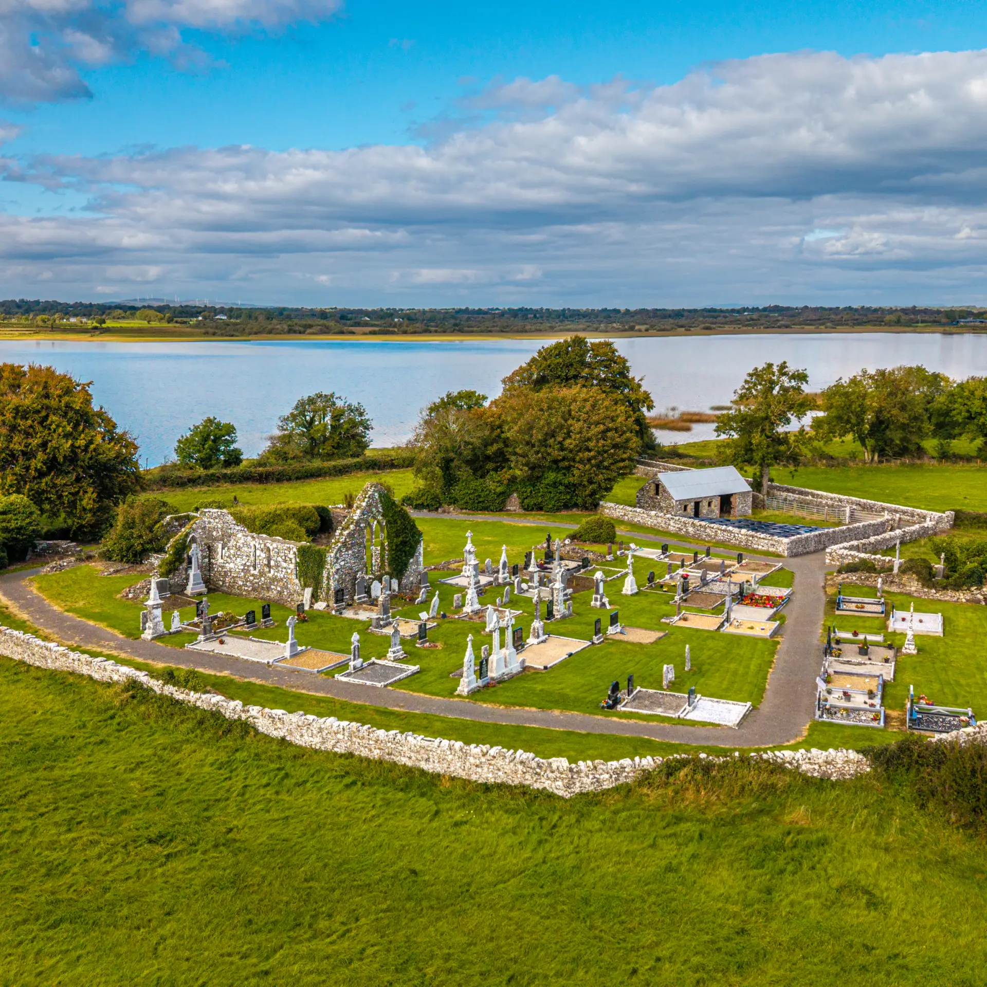 Aerial view of an old stone church ruin and graveyard surrounded by green fields, near a lake under a partly cloudy sky, with trees and hedges bordering the cemetery.