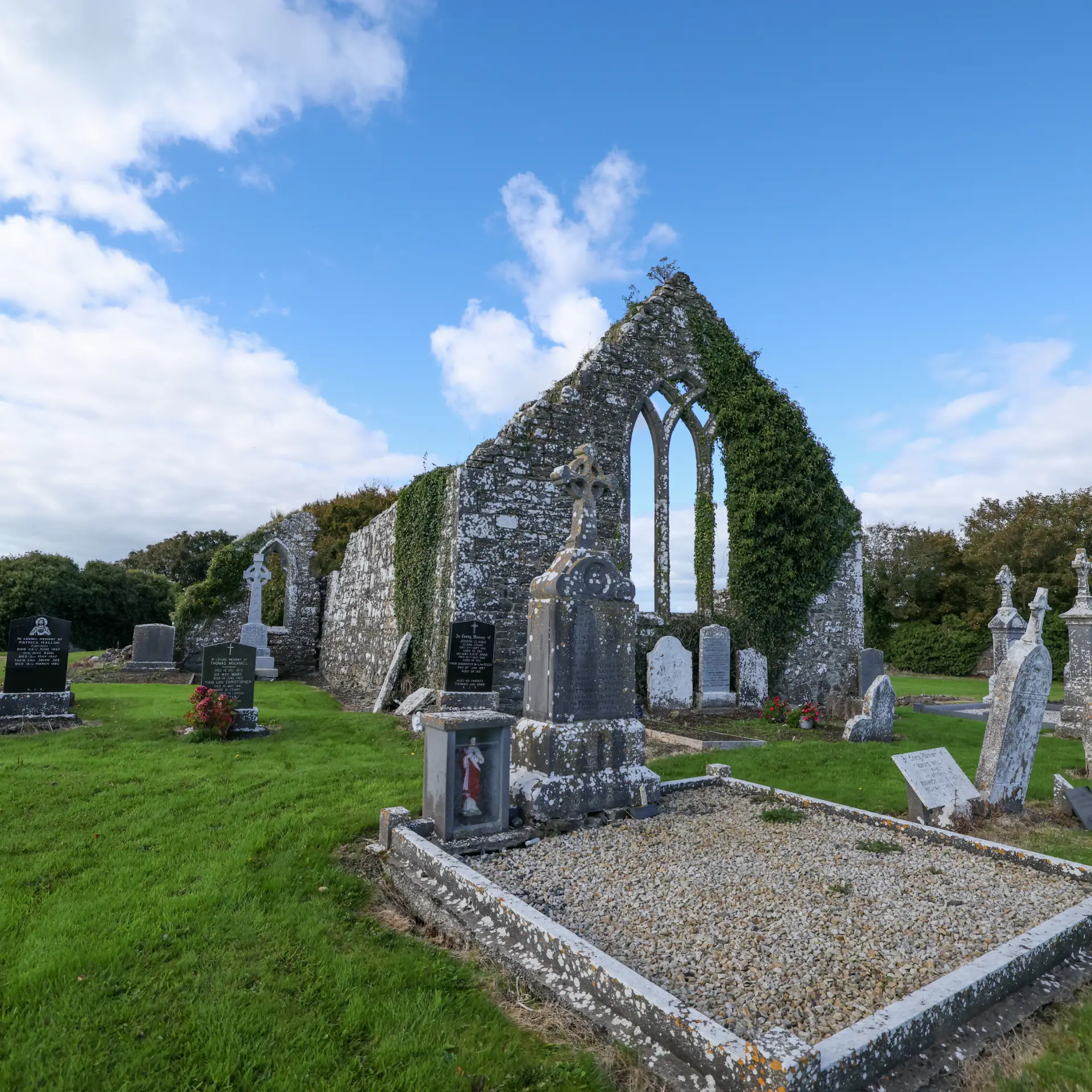Old stone church ruins with ivy-covered walls stand in a graveyard filled with weathered headstones and green grass under a bright blue sky with scattered clouds.