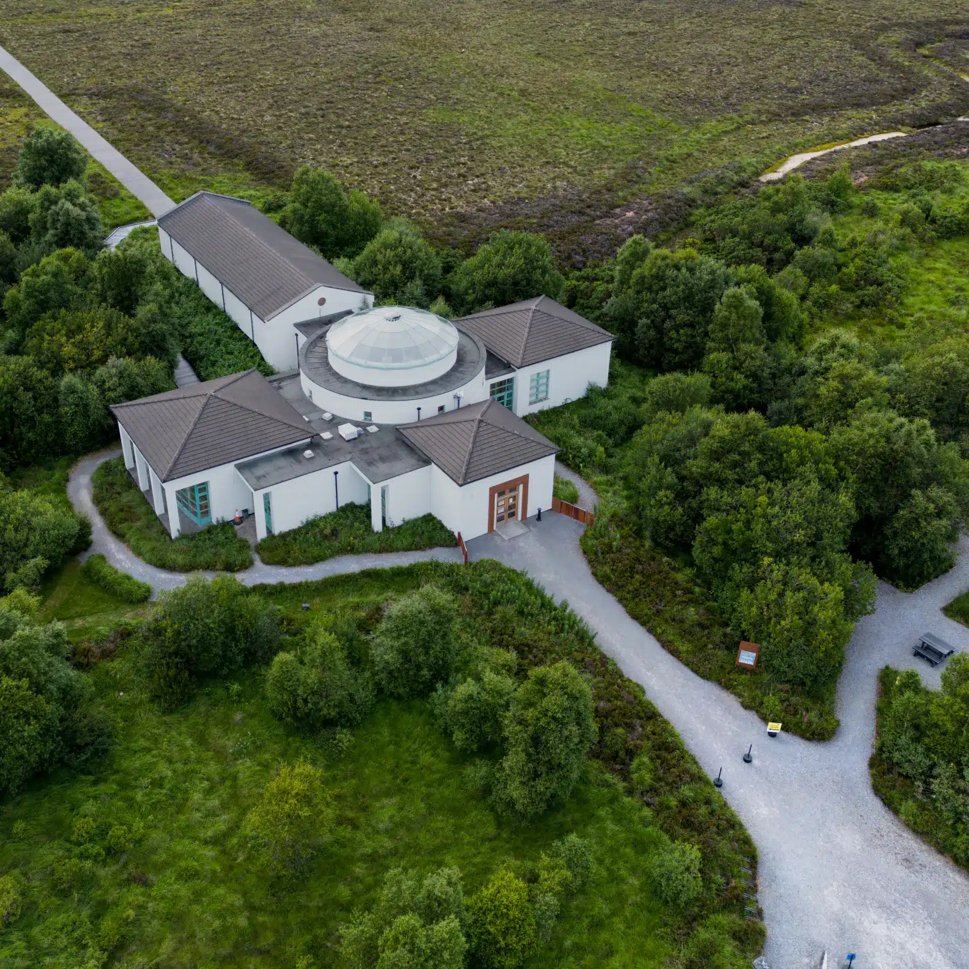 Aerial view of a white, modern building with a central glass dome, surrounded by green trees, walking paths, and open grassy land. The area looks peaceful and well-maintained.