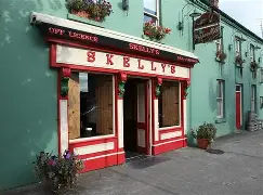 A green building with red and white trim features a pub called Skelly's. The entrance has wooden doors, flower pots, and hanging plants, with “Off Licence” and “Lounge” signs above the door.