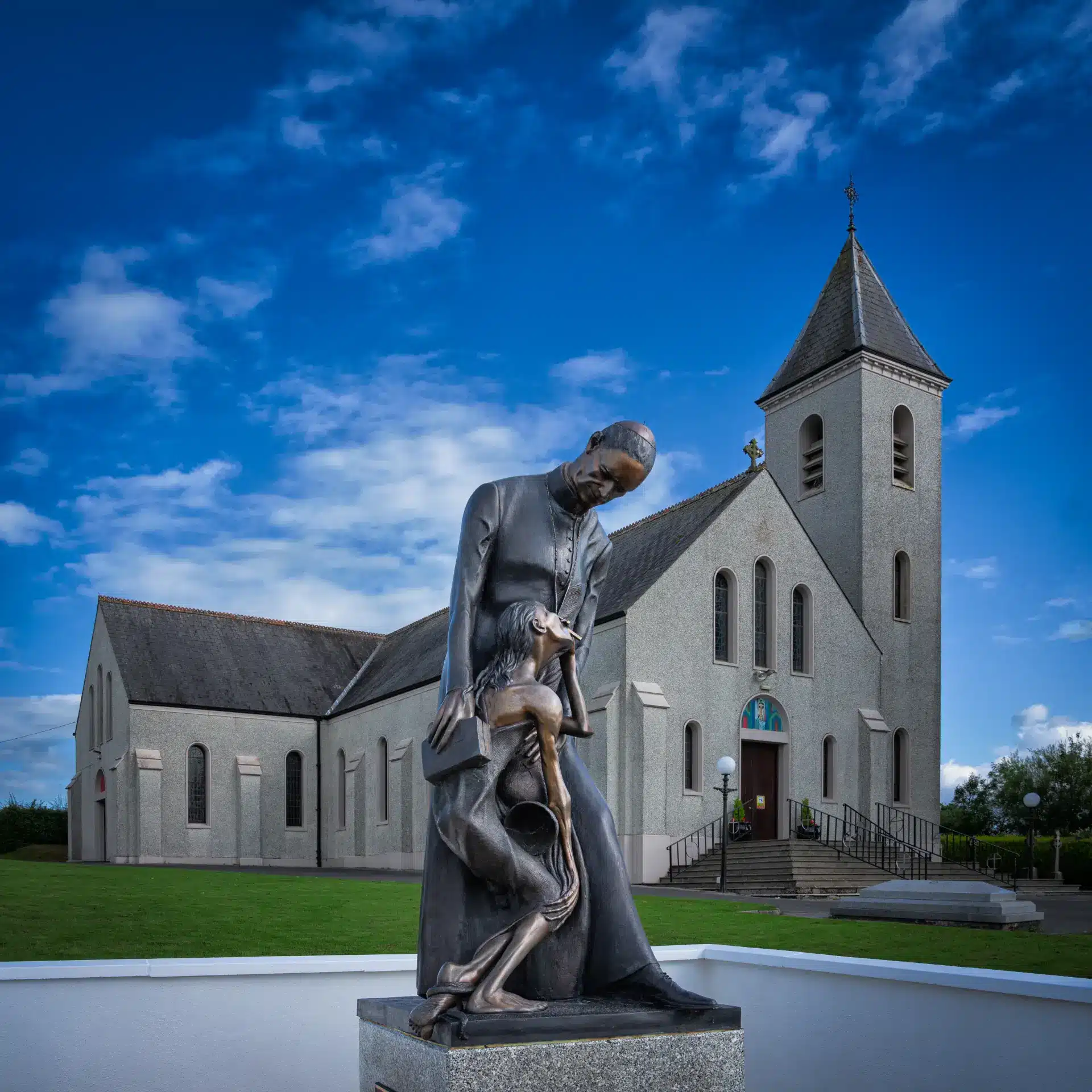 A bronze statue of a man embracing a child stands in front of a grey stone church with a tall spire, under a blue sky with scattered clouds. Grass and trees surround the church.