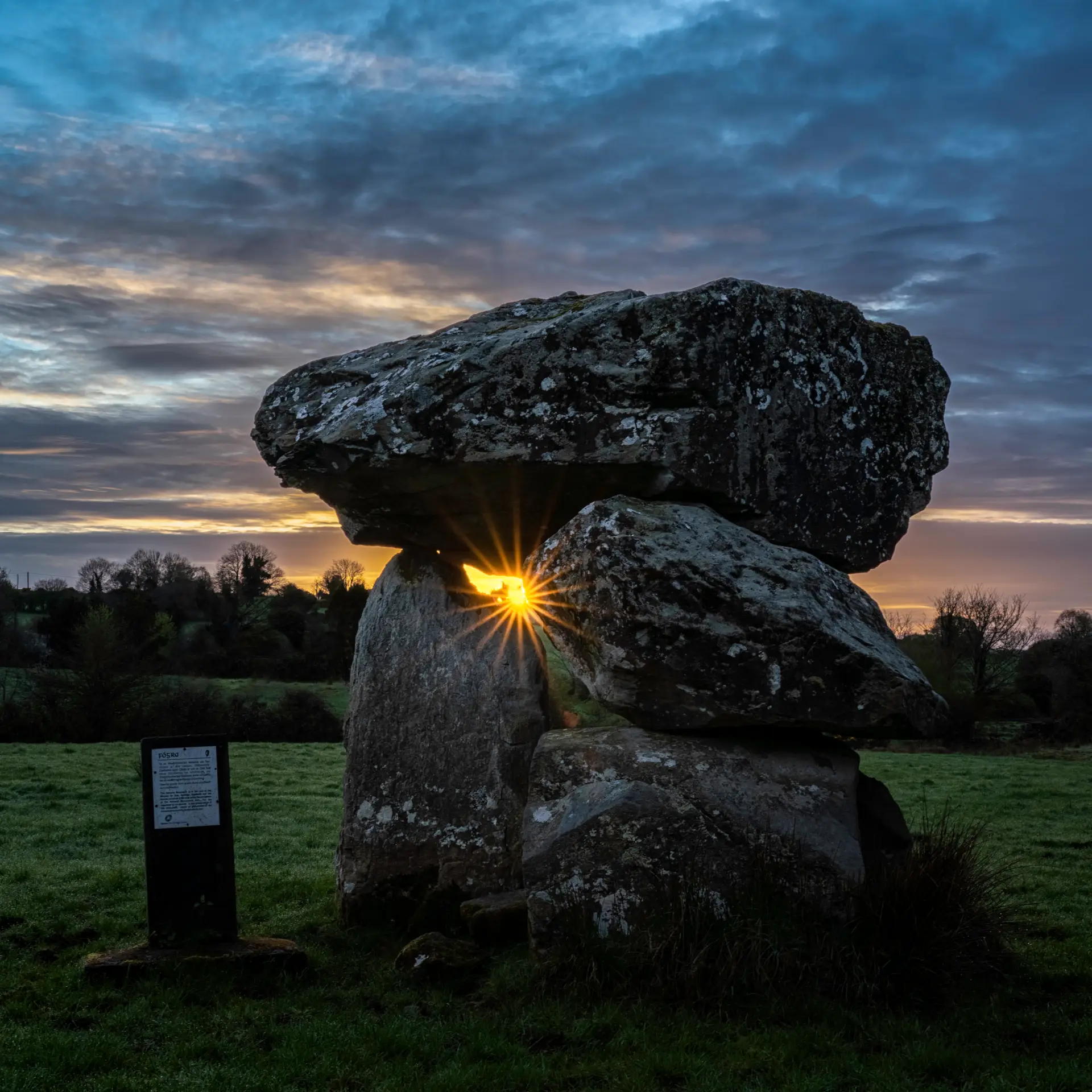 A large ancient stone structure stands in a grassy field at sunrise, with sunlight shining through a small gap between the stones. There is a plaque on a post nearby. Trees and a dramatic cloudy sky are in the background.